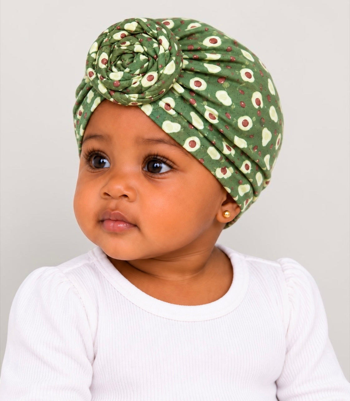 A baby wearing a green floral turban headband with a large rose detail on top, paired with a white ribbed long-sleeve top, photographed against a light gray background. Fatisha Boutique